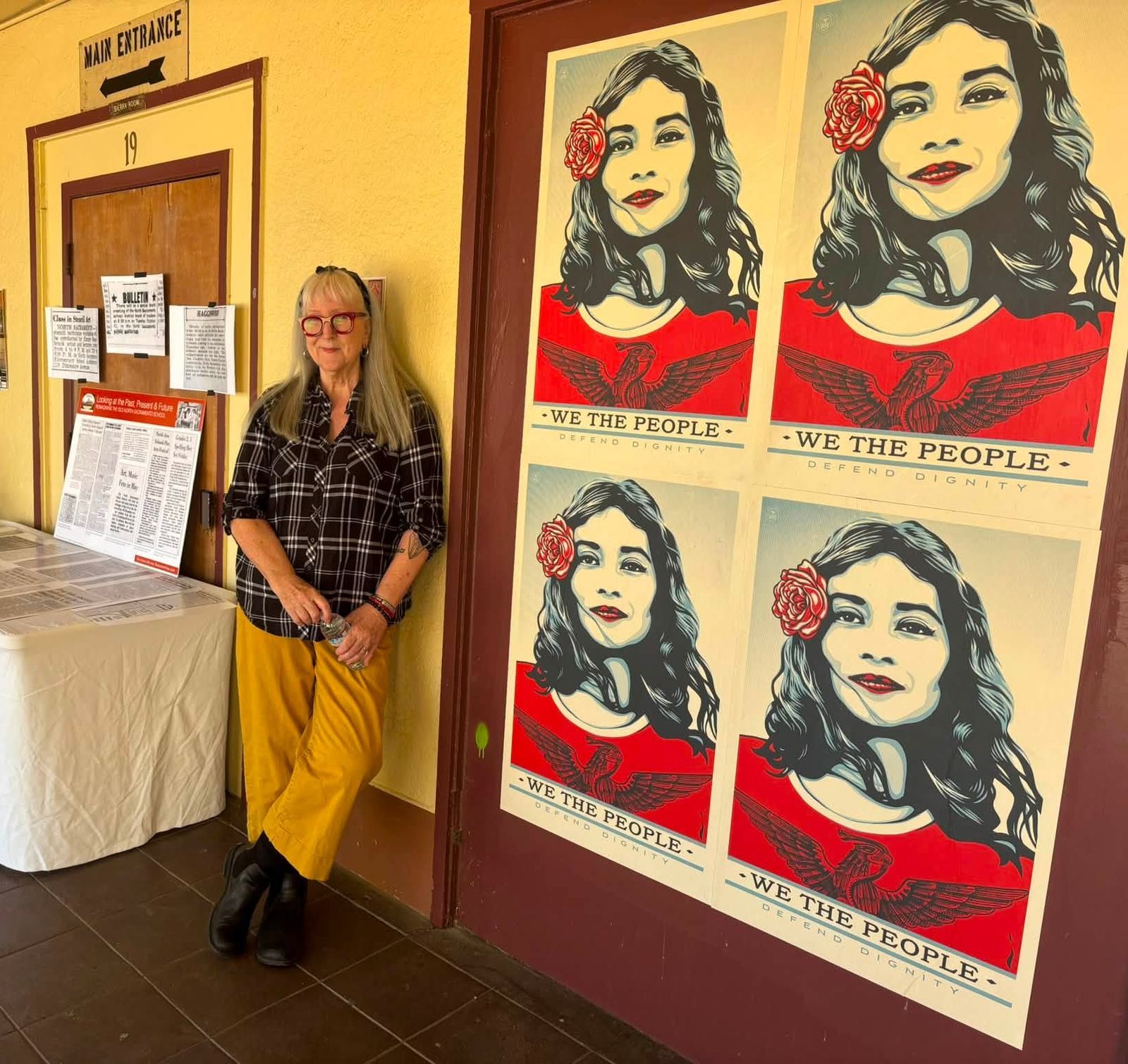 Four Shepard Fairey We the People posters on a door beside the school's main entrance, with a history table and a visitor nearby